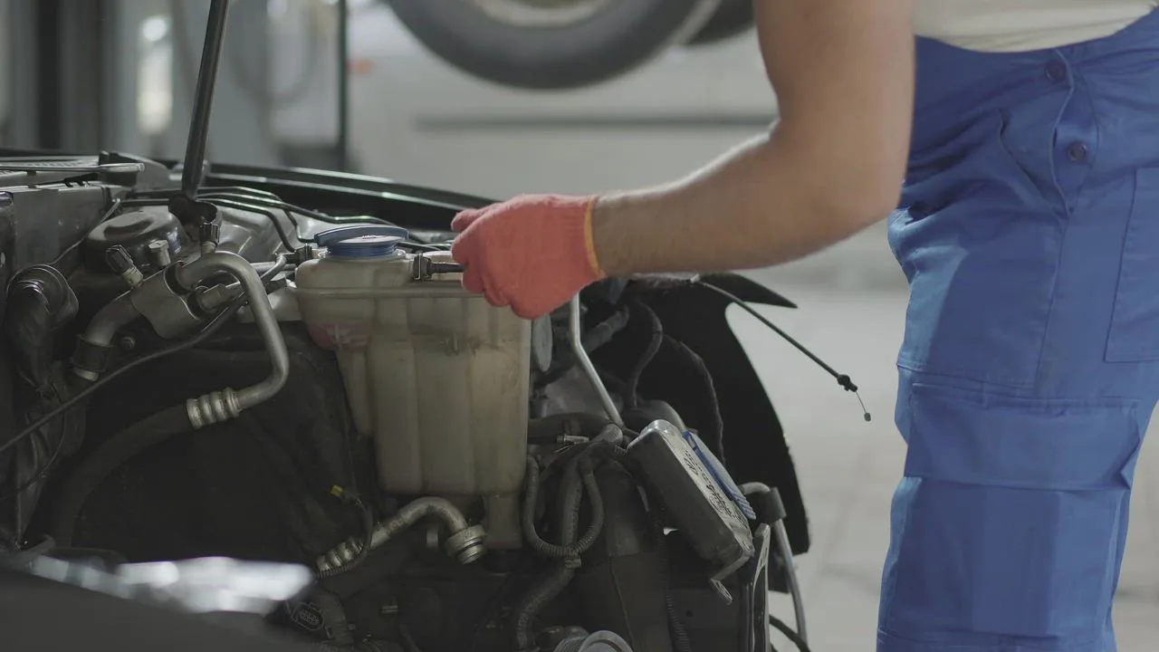 Gloved hand at the coolant reservoir beside the A/C lines in an engine bay
